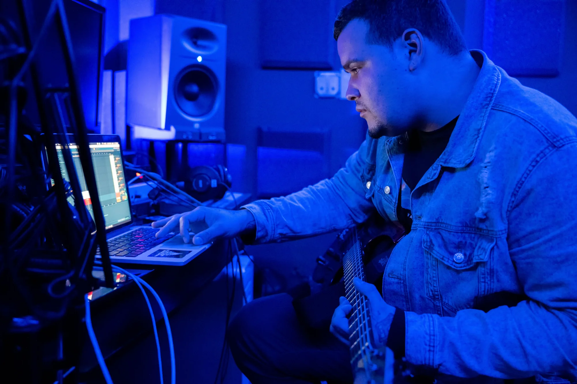A guitarist working with a laptop in a low-lit recording studio
